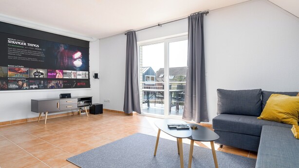 A modern living room with a grey sofa, a small black coffee table, and a TV mounted on the wall displaying a Netflix original series. - 90 sqm Cozy home cinema with balcony & projector (Kassel)