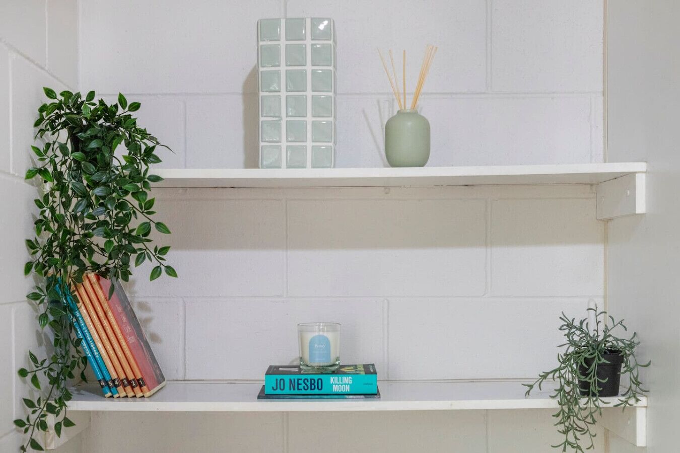 A white tiled wall with three shelves. The top shelf has a white vase with reed sticks, the middle shelf has a small glass candle, and the bottom shelf has a book titled 'JO NESBO KILLING MOON'.