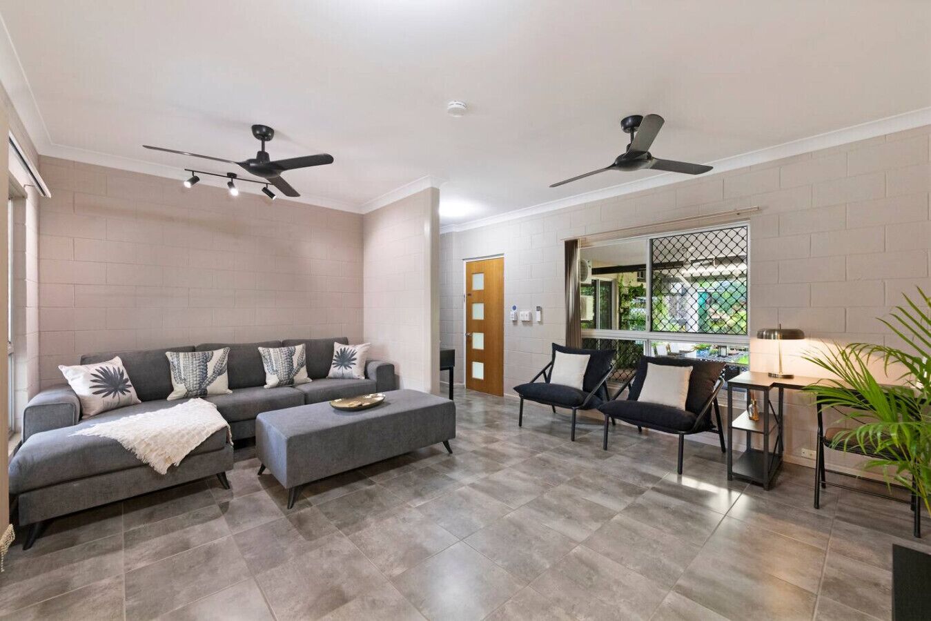 A modern living room with a grey sofa, two armchairs, a coffee table, and a dining area with a table and chairs. There are ceiling fans, a large window with a view, and a door leading to another room.