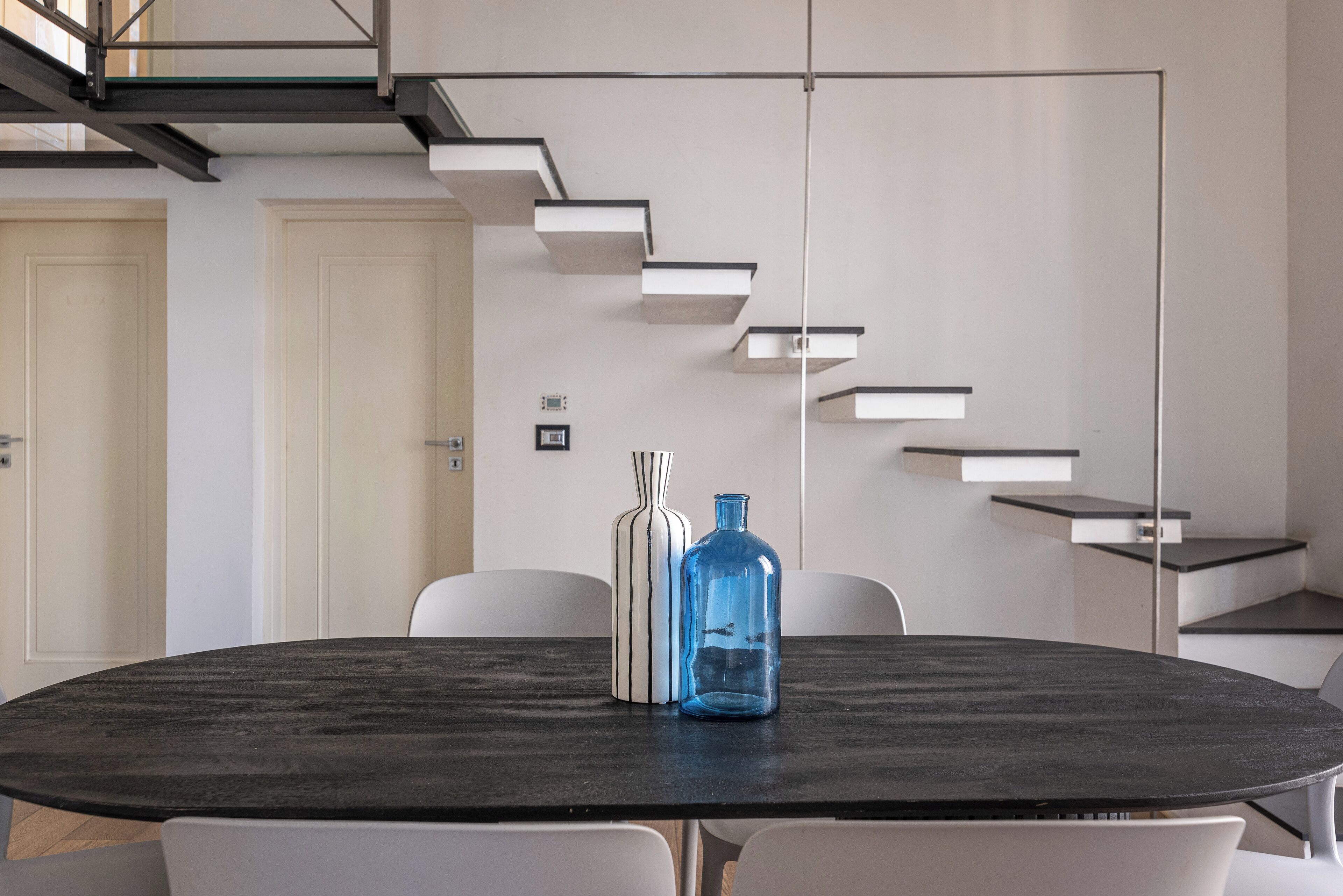 Una sala de conferencias moderna con una mesa de mármol oscuro, sillas blancas, un jarrón azul y un jarrón blanco. Se ve una escalera con estantes empotrados al fondo.