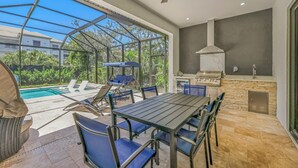 Un patio exterior con mesa y sillas, una parrilla y vistas a una piscina y zonas verdes.