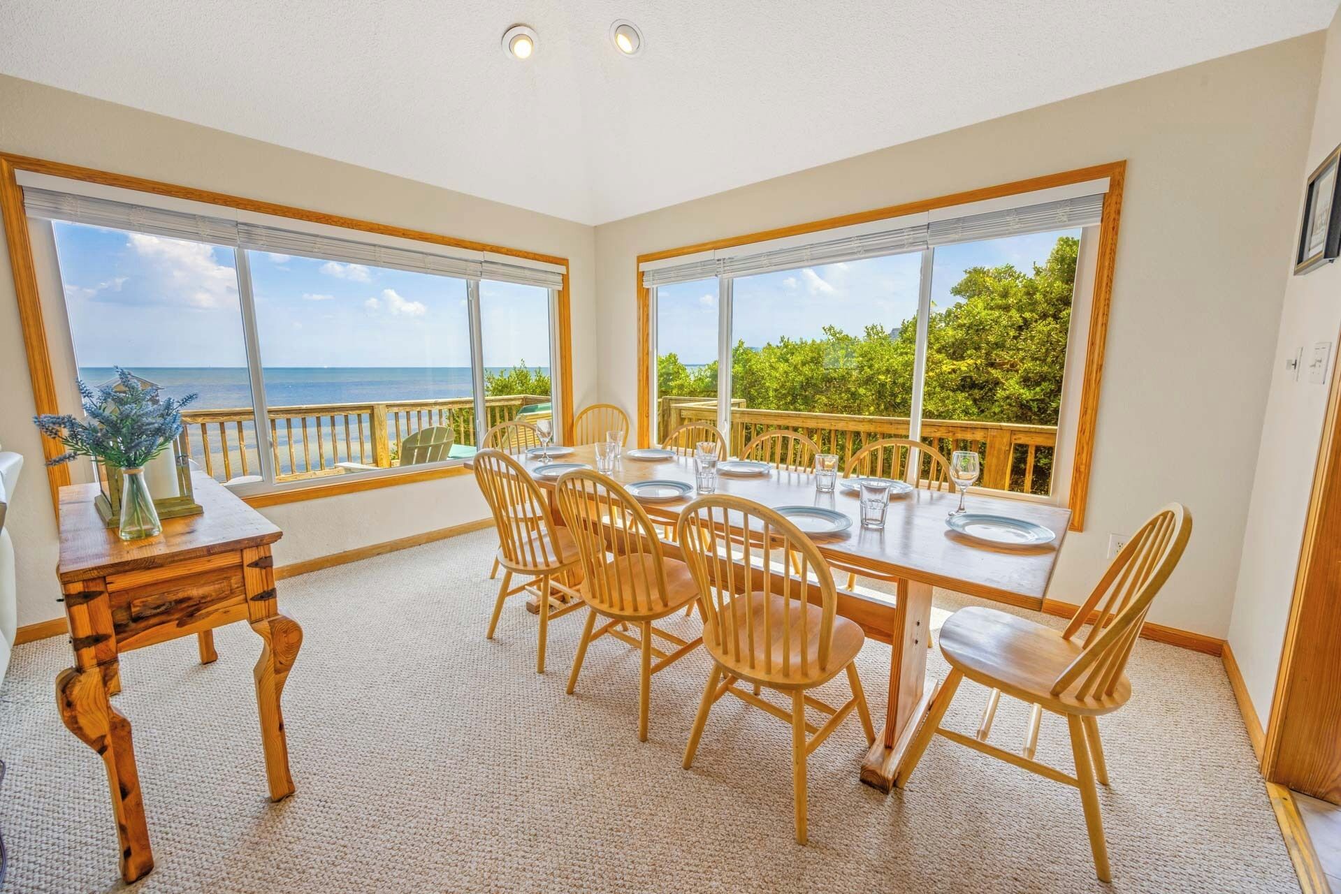 A dining area with a table set for six, large windows with a view of the ocean, and a wooden deck outside.
