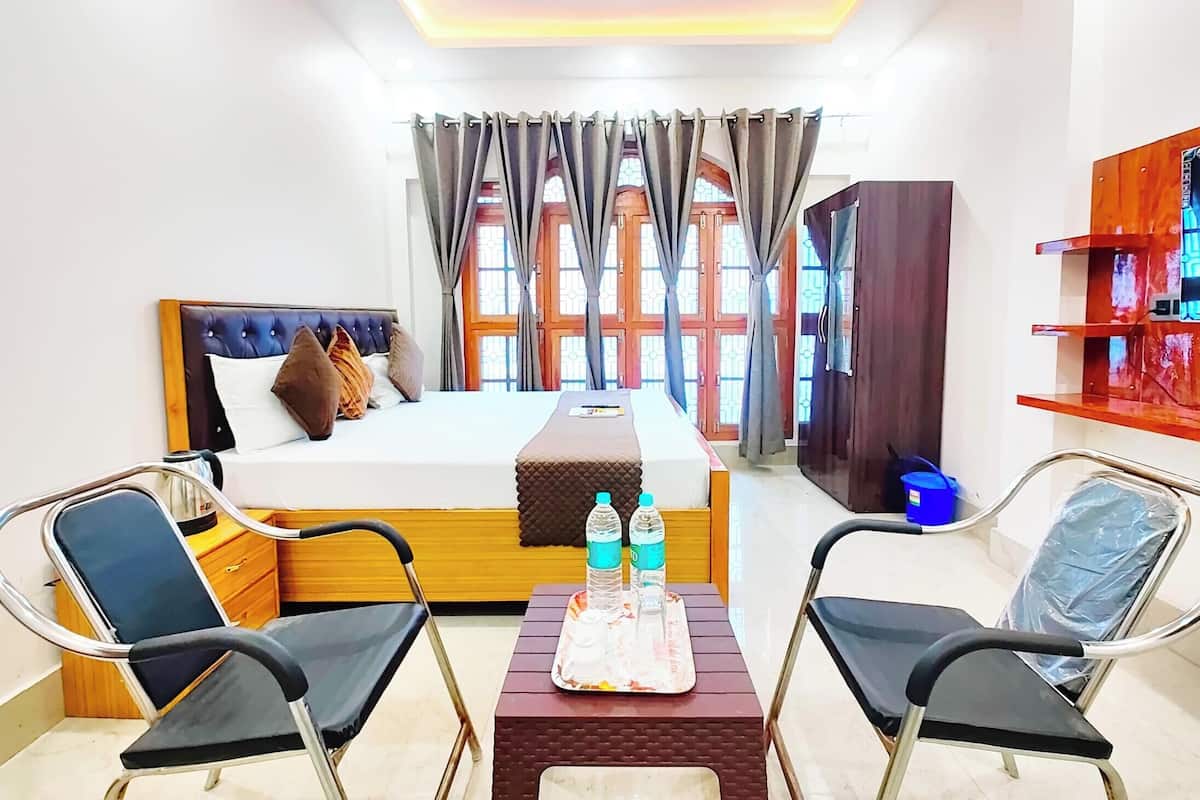 Quarto de hotel com cama, duas cadeiras, uma mesinha com garrafas de água, um armário de madeira e uma janela com cortinas.