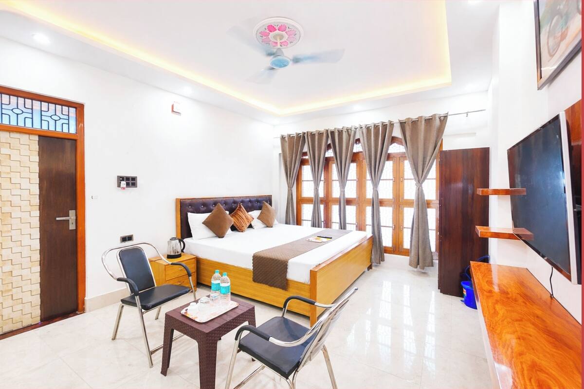 Quarto de hotel com cama, escrivaninha, cadeiras, televisão e ventoinha no teto.