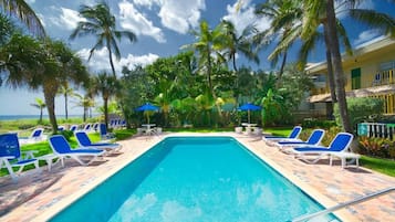 Ein Swimmingpool mit Liegestühlen und Sonnenschirmen, umgeben von Palmen und einem Gebäude mit Balkonen.