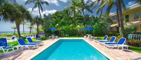 A swimming pool with lounge chairs and umbrellas, surrounded by palm trees and a building with balconies.