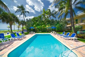 Una piscina con sillones de playa y sombrillas, rodeada de palmeras y un edificio con balcones.