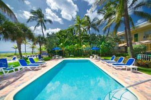 Una piscina con sillones de playa y sombrillas, rodeada de palmeras y un edificio con balcones.