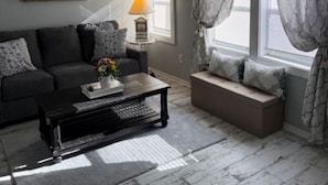 A living room with a dark sofa, a coffee table, and a window with patterned curtains. - Storybook Chatham (Newport)