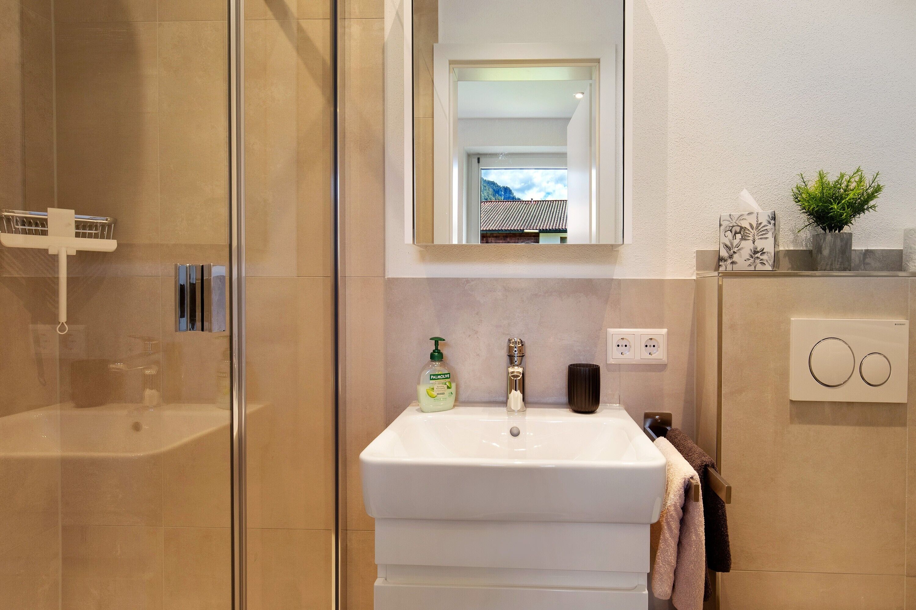 A bathroom with a glass shower enclosure, a white sink with a mirror above, a bottle of hand soap, and a potted plant.