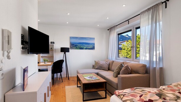 Una camera moderna con TV a schermo piatto, divano, letto, scrivania con sedia e finestra vista montagna.
