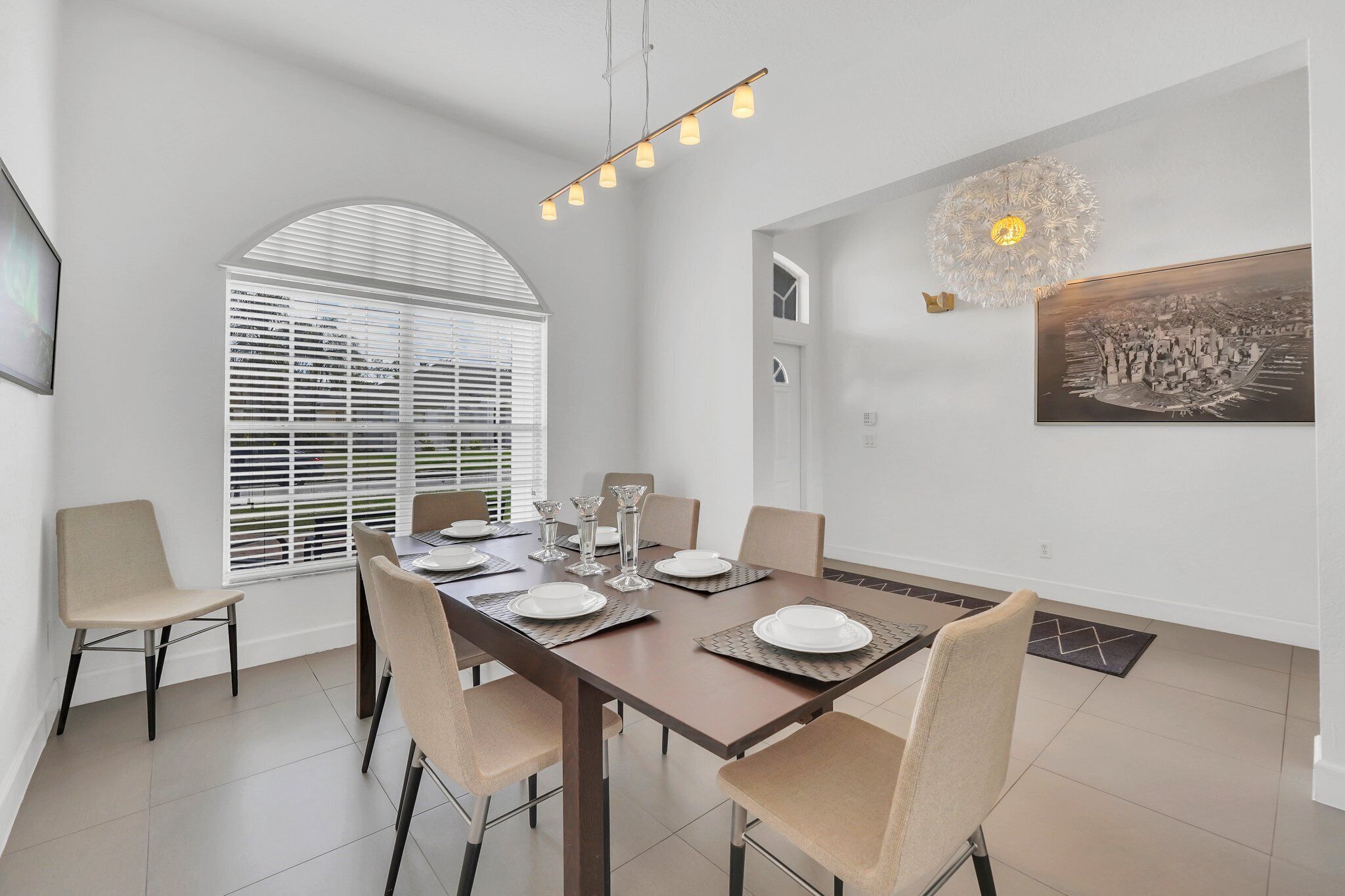 Een eethoek met een lange tafel gedekt voor vier personen, omringd door stoelen, met een groot raam en een decoratieve plafondlamp.