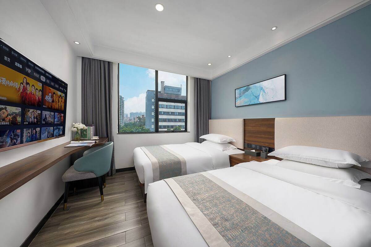 A hotel room with two beds, a desk, a chair, a large window with a city view, and a wall with movie posters.