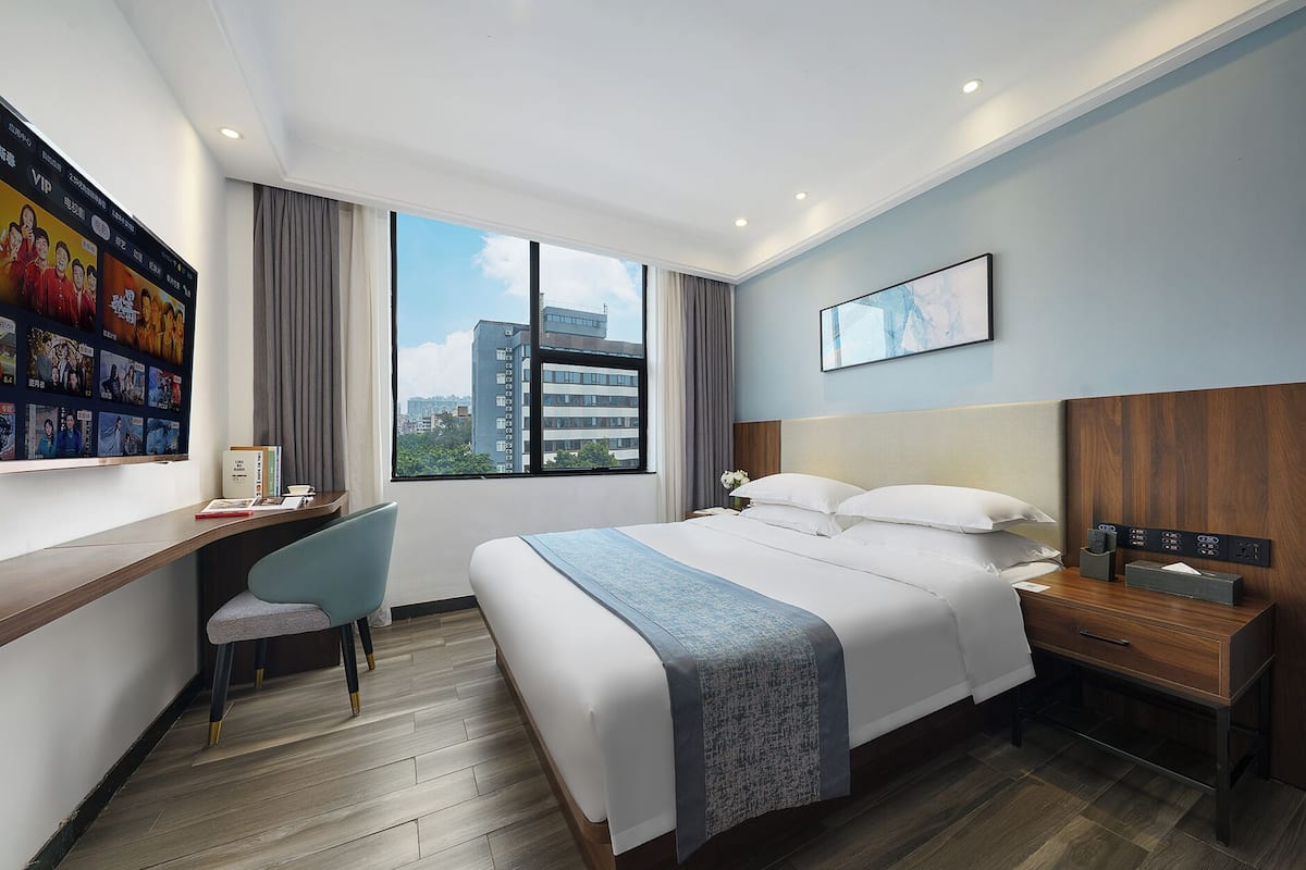 A hotel room with a bed, bedside table, desk, chair, and a large window offering a city view.