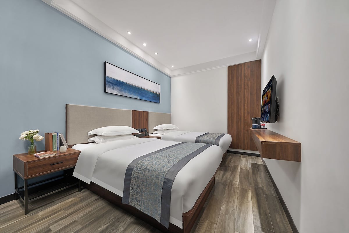 A modern hotel room with two beds, a bedside table with a lamp, a mounted TV, and a painting on the wall.