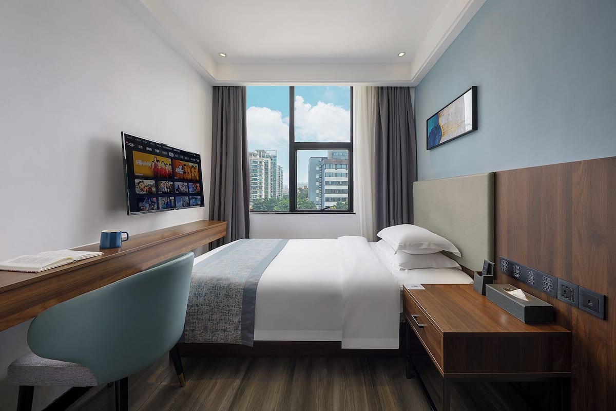A hotel room with a bed, a desk, a chair, a TV mounted on the wall, and a window with a view of buildings.