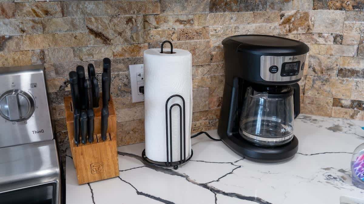 Una cocina con protector contra salpicaduras de piedra, cafetera, dispensador de toallas de papel y bloque de cuchillos.