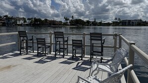 Une terrasse en bois, agrémentée de chaises et de tables, qui offre une vue sur une étendue d’eau, avec des bâtiments et des palmiers en arrière-plan.