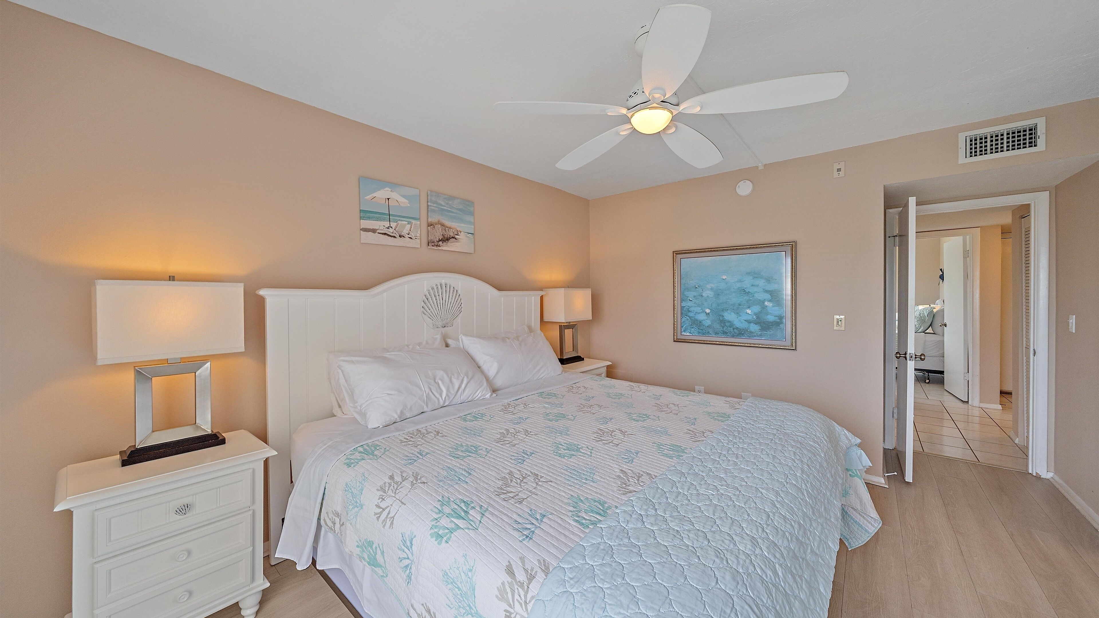 A bedroom with a bed, two bedside tables with lamps, a ceiling fan, and artwork on the walls.