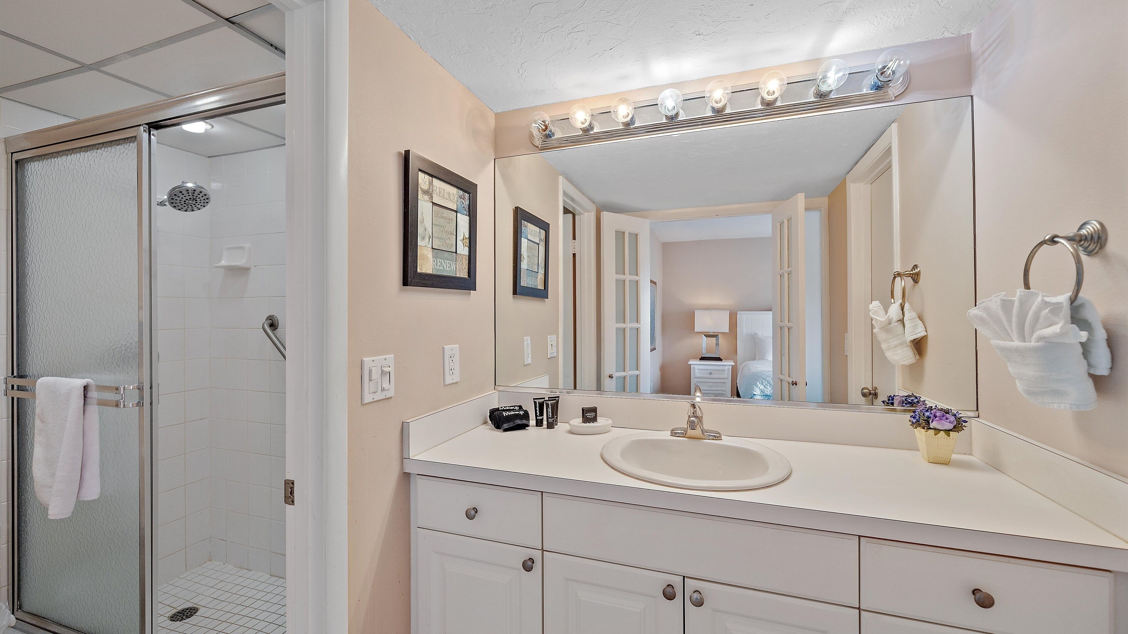 A bathroom with a glass shower enclosure, a vanity with a sink, and a large mirror with lights above.
