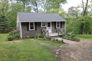 A small, single-story house with a porch, surrounded by a garden and trees. - Cozy Cottage by Molls Pond  2409 (Eastham)