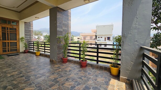A covered balcony with stone flooring, potted plants, and a view of surrounding buildings. - Bora's Homestay (Haldwani)