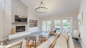 A spacious living room with a fireplace, a large TV, wicker furniture, and a balcony with outdoor seating. - Private home in Hilton Head Island with a private pool and hot tub! (Hilton Head)