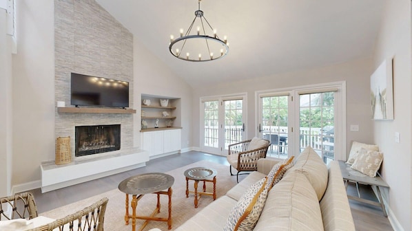 A spacious living room with a fireplace, a large TV, wicker furniture, and a balcony with outdoor seating.