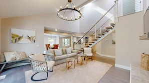 A spacious living area with a high ceiling, a staircase with a glass railing, and a chandelier.
