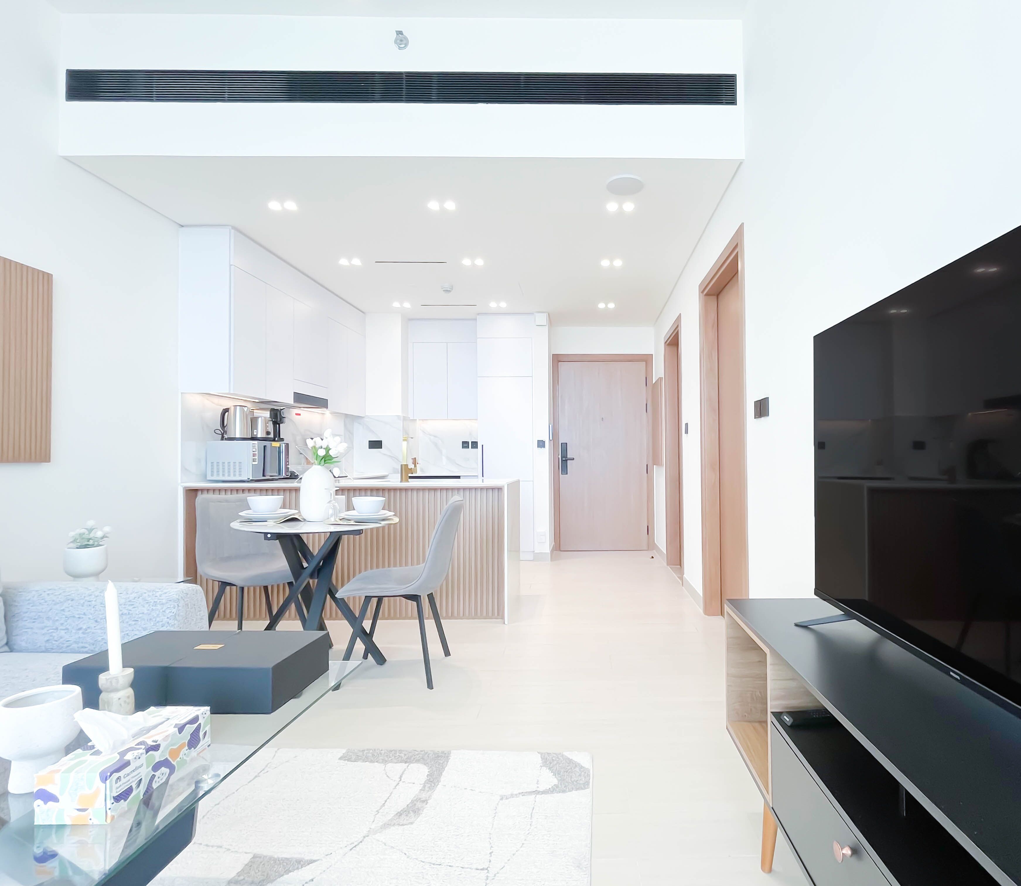 A modern living room with a flat-screen TV, a dining area with a round table and chairs, and a kitchen in the background.