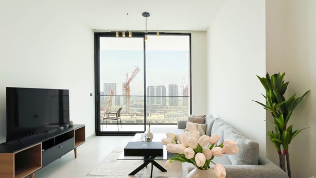 A modern living room with a large flat-screen TV, a grey sofa, a dining table with a vase of flowers, and a view of a cityscape through a large window. - Luxury & Comfort 1B at Binghatti Amber & Balcony (Dubai)