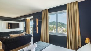 A modern hotel room with a large window, a black sofa, a flat-screen TV, and a bed with a white comforter.