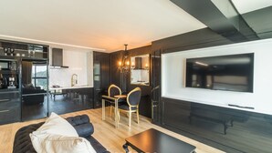A modern living room with a sofa, a coffee table, and a dining area with chairs and a table. A large flat-screen TV is mounted on the wall.