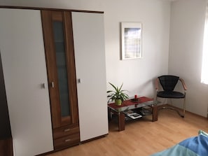 A room with a wooden wardrobe, a glass-top table with a plant, a black chair, and a framed picture on the wall.