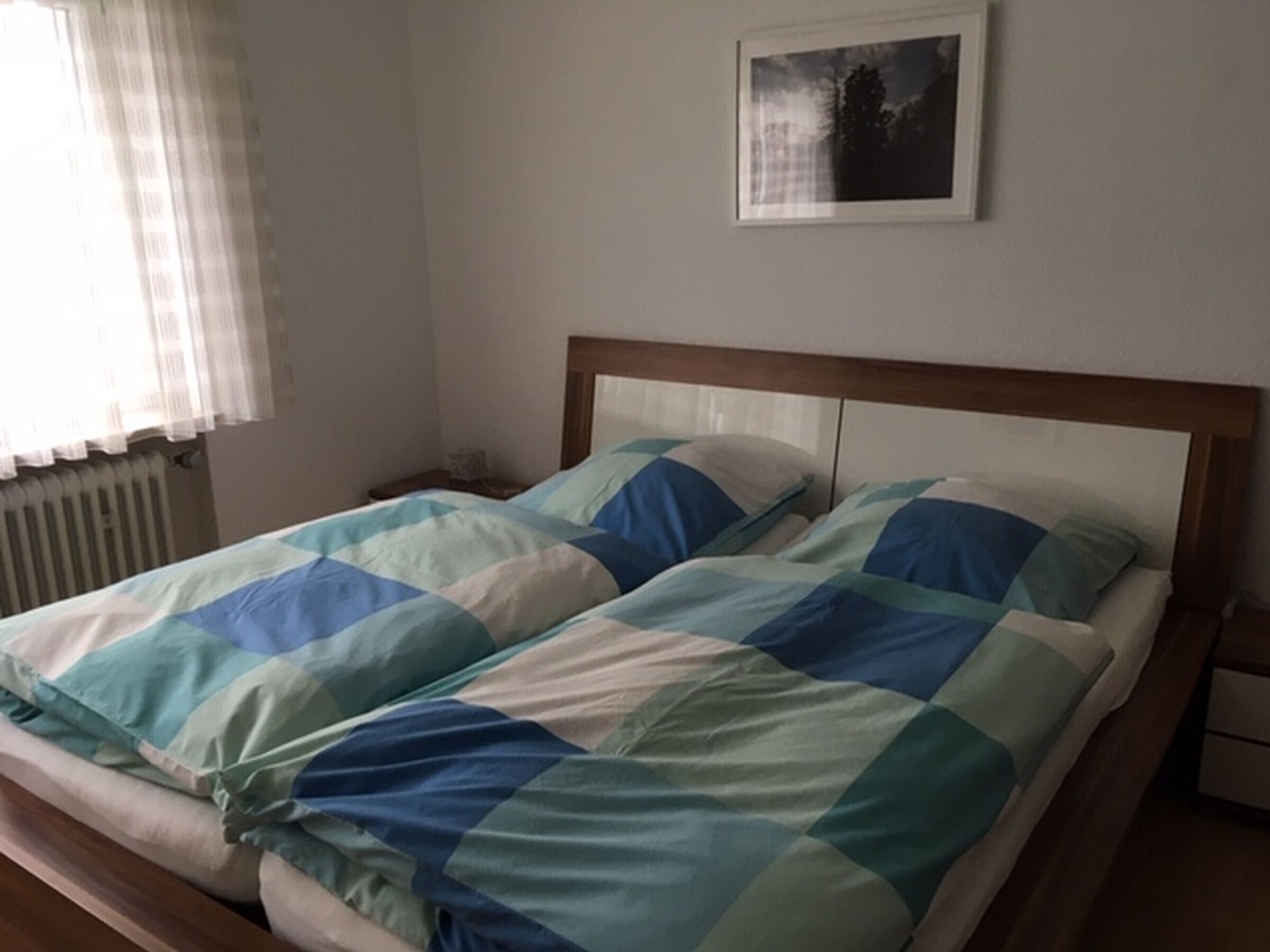 Un lit avec une couvre-lit à carreaux, une table de chevet, une commode, un tableau encadré au mur et une fenêtre avec des rideaux.