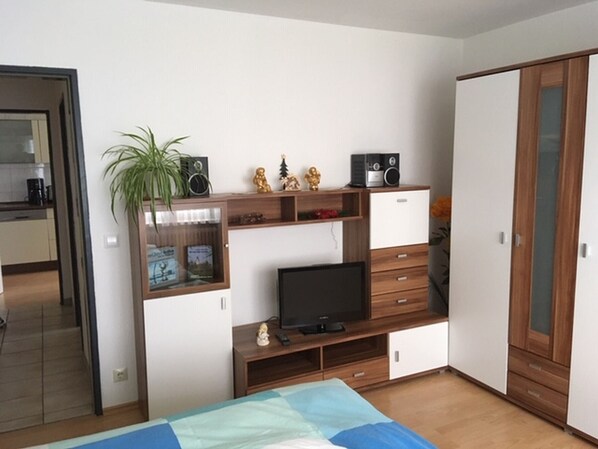 A bedroom with a bed, a TV, a wardrobe, a small cabinet with a plant, and a kitchen area visible through an open door.
