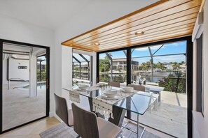 A modern dining area with a glass table and chairs, large windows offering a view of a marina and buildings, and a wooden ceiling.