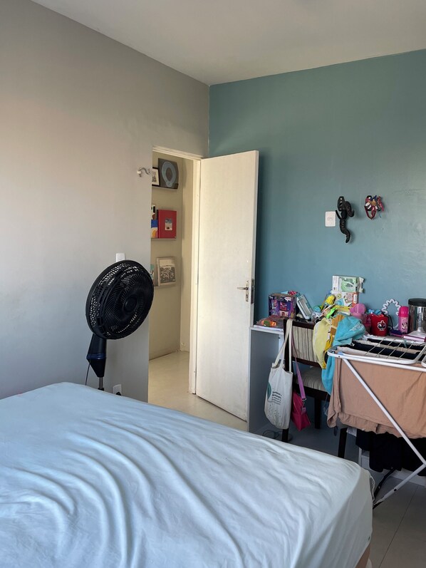 Une chambre à coucher avec un lit, un ventilateur sur pied, une porte donnant sur une autre pièce et une table encombrée d’objets divers.