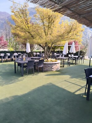 Area esterna con tavoli e sedie, circondata da alberi e un vaso in pietra.