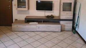 Un salon avec un téléviseur à écran plat fixé à un mur en bois, une petite étagère ornée d’objets décoratifs et une porte équipée d’une caméra de surveillance.