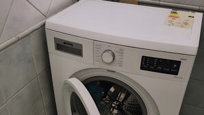 Une machine à laver à chargement frontal, placée dans un coin d’une pièce dont les murs et le sol sont recouverts de carreaux.