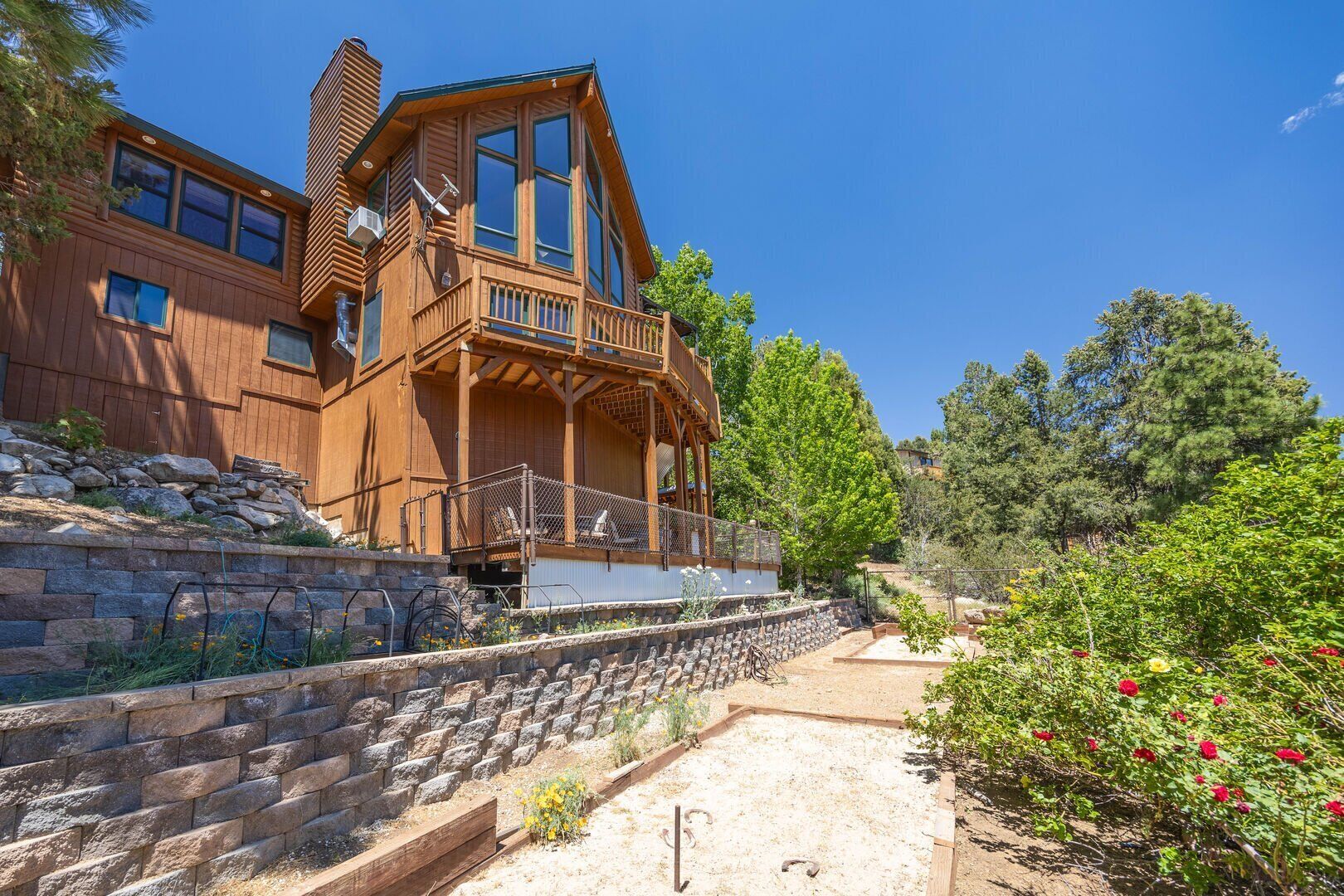 A two-story wooden house with a balcony, stone retaining wall, and a garden with flowers.