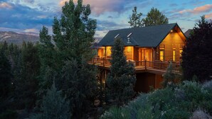 Une cabane en rondins dotée d’un balcon, entourée d’arbres et avec une chaîne de montagnes en arrière-plan.