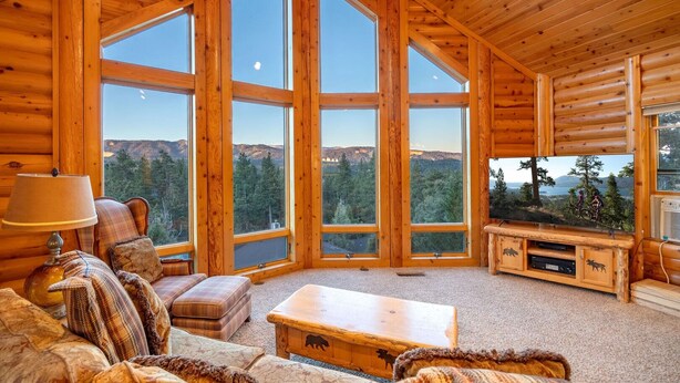L’intérieur d’une cabane en rondins, avec de grandes fenêtres, une cheminée et un coin salon confortable.