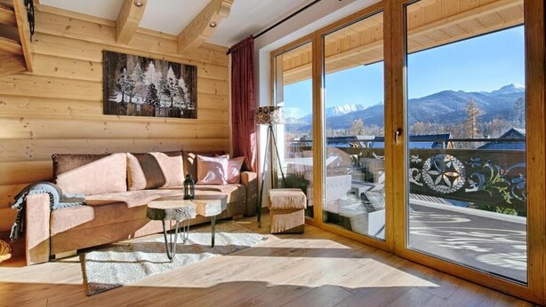 Ein gemütliches Blockhaus-Interieur mit Holzboden, einer Couch, einem kleinen Tisch und Blick auf Berge durch große Glasüren.