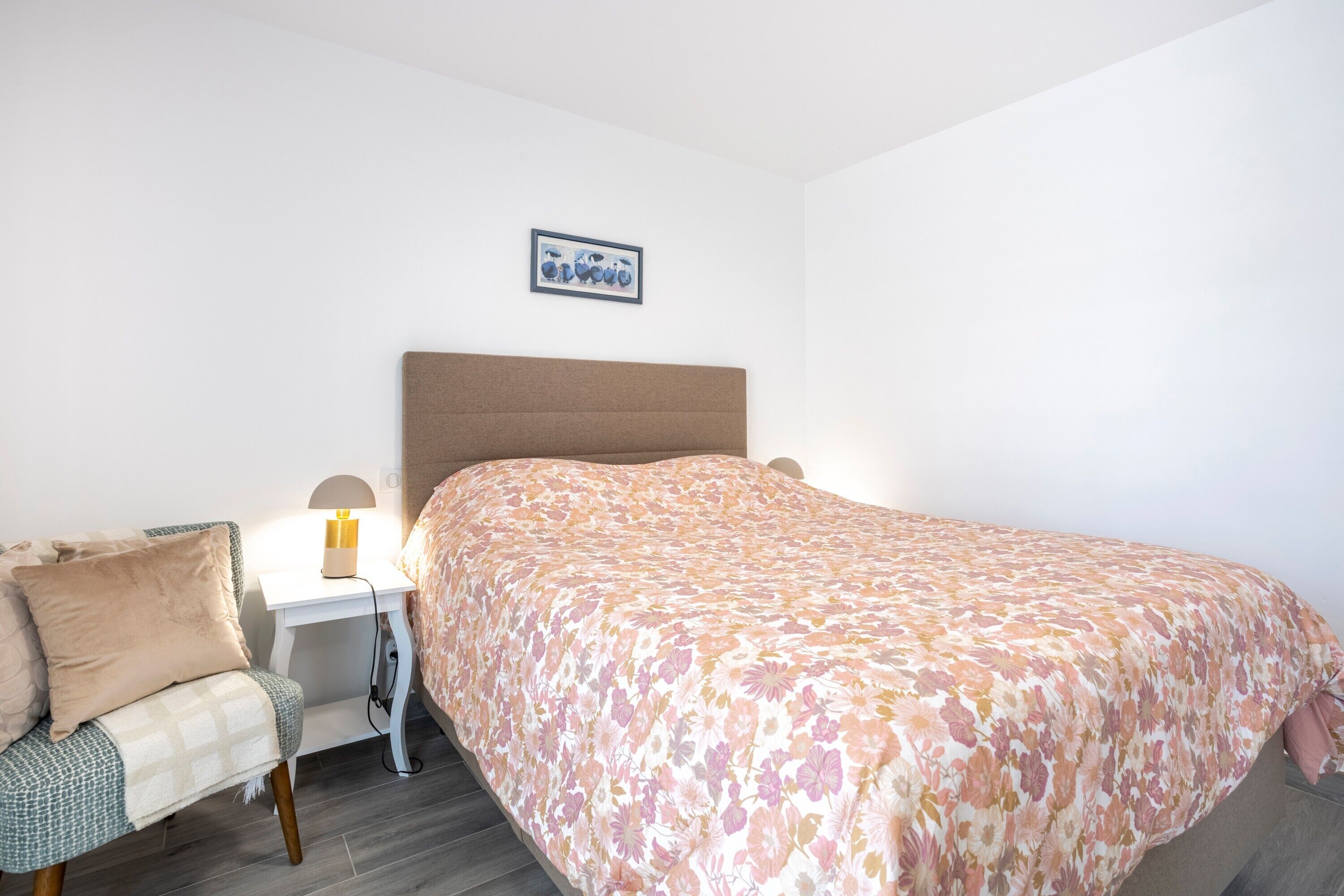 A bedroom with a bed, bedside table, lamp, chair, and a framed picture on the wall.