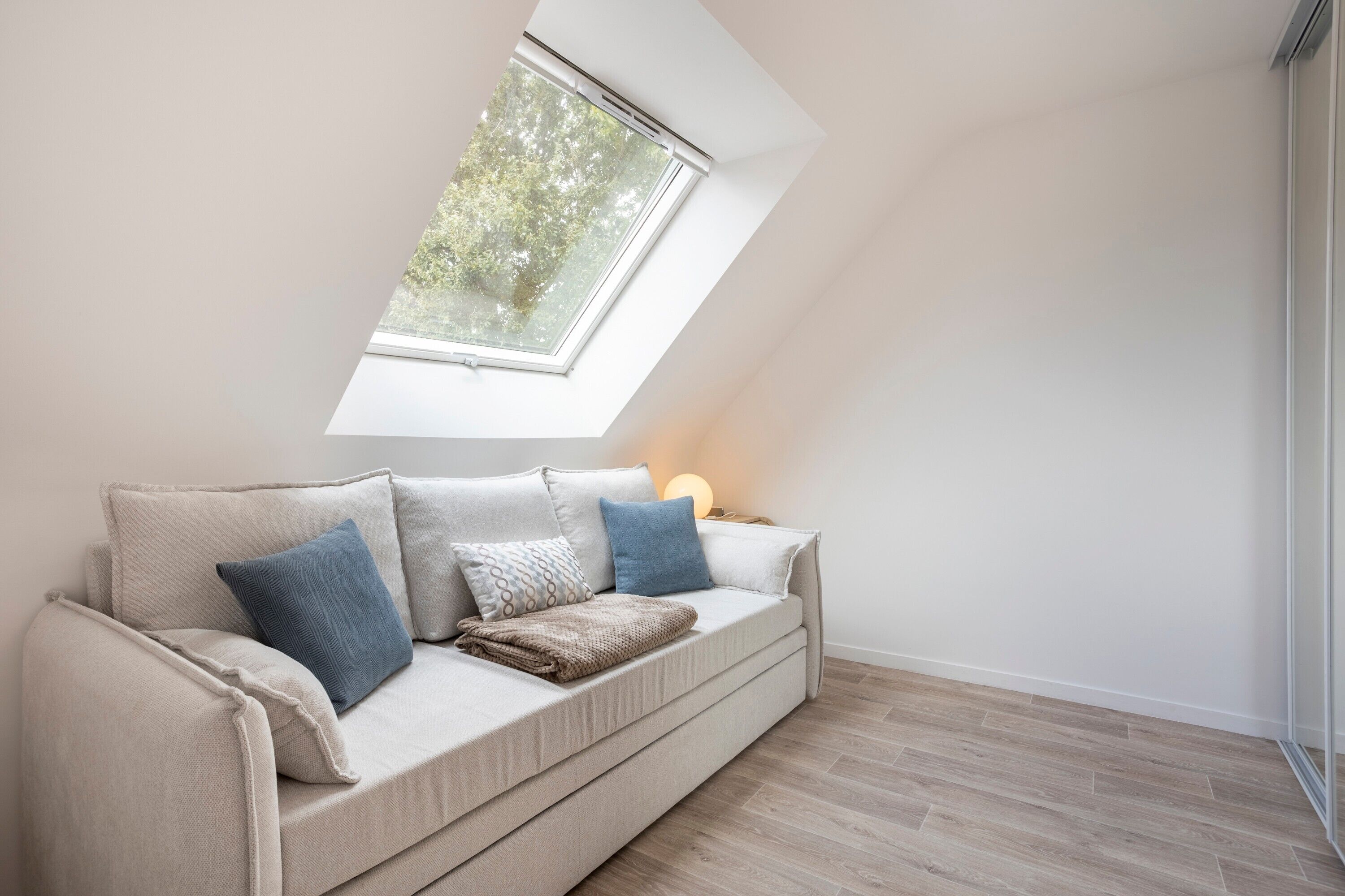 A room with a sloped ceiling, a sofa with cushions, a window, and a lamp.