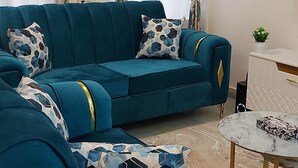 A teal sectional sofa with patterned cushions, a small round table with a marble top, and a potted plant on a white side table.