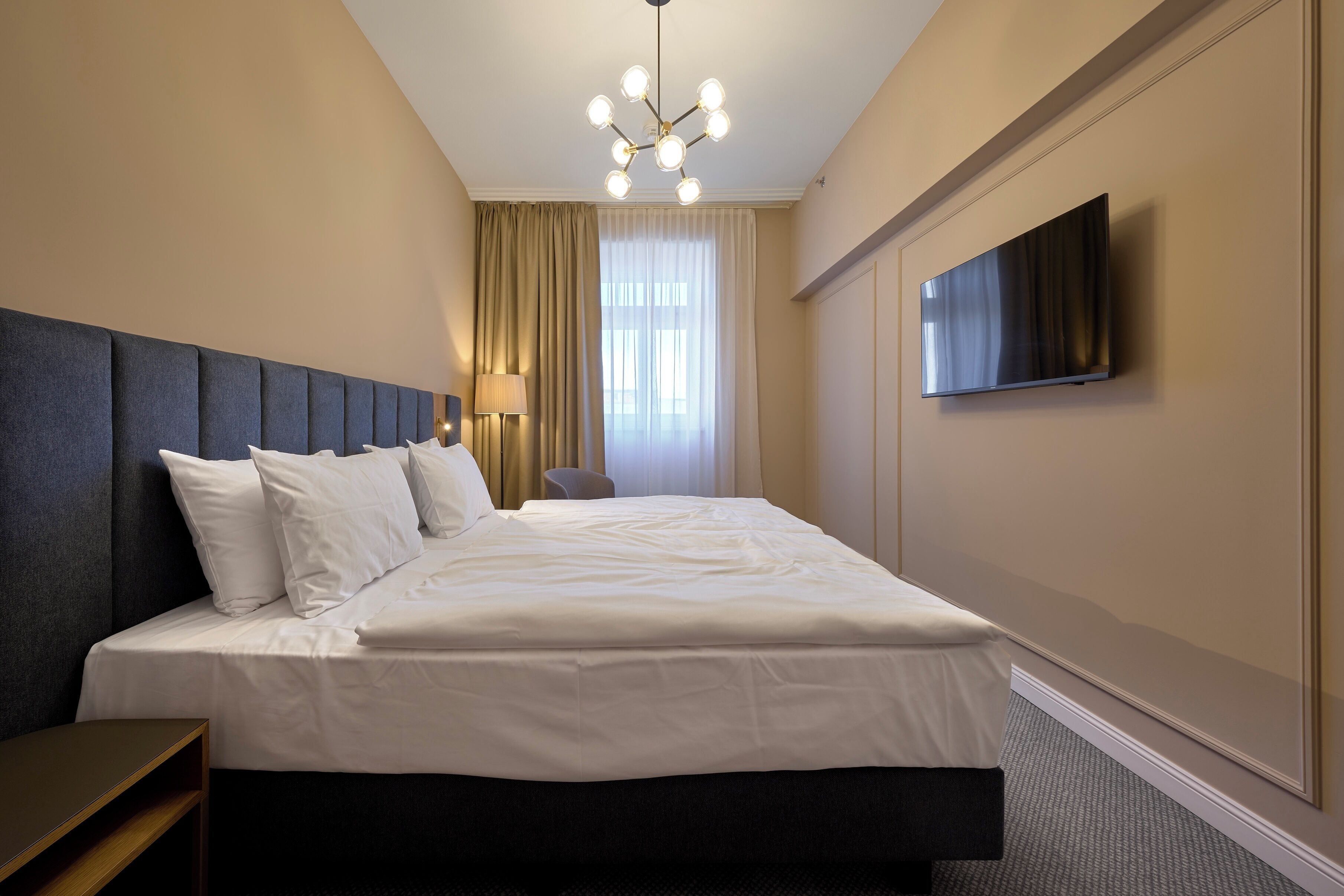 Quarto de hotel com uma cama grande, televisão de tela plana na parede, luminária no teto e janela com cortinas.