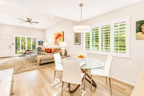 A modern living room with a glass dining table, white chairs, a sofa, and a ceiling fan.